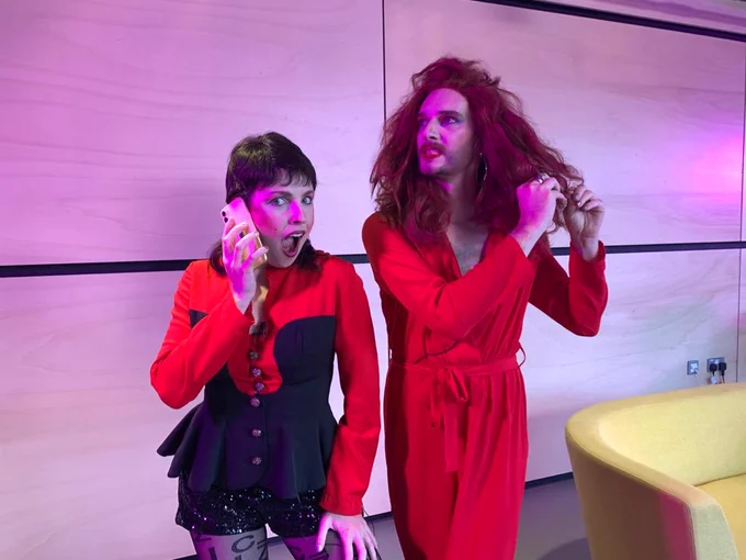 TomYumSim stand waiting to go live in the studio. Tom is wearing a red jumpsuit, a curly red wig and red lipstick. Simone holds a phone and is wearing a red and black peplum top, patterned tights and a brown mullet wig.