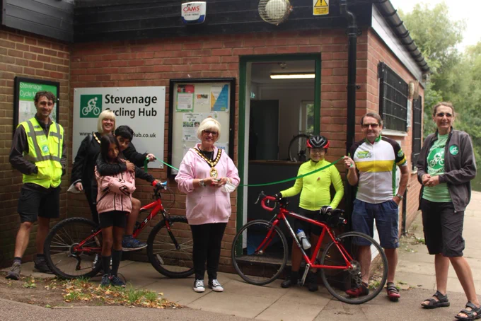Official opening with a group of people standing outside the open Hub building.  People left to right, cycle instructor in high vis, Mayoress with chain of office, two young cyclists in front in a cute cuddle and a red bike, Mayor Councillor Sandra Barr with chain of office,  a child with another red bike, Councillor Richard Henry in Cycling Festival top, and Tina in Cycling Hub branded clothes.  Mayoress and Councillor Henry holding the ribbon for the Mayor to cut.