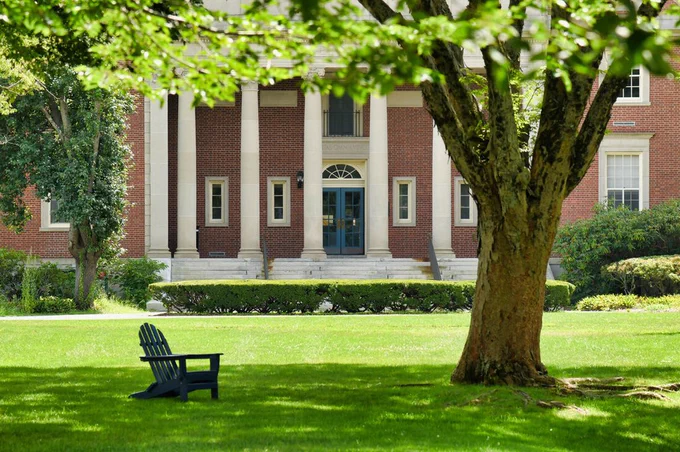 Brick building with ivory columns framed by a giant tree on the right. Blue Adirondack chair on lush green grass in front of the building. This photo was taken on a sunny day.
