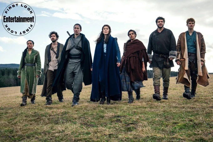 A group of adventurers, Nynaeve al'Meara (Zoë Robins), Matrim Cauthorn (Barney Harris), Lan Mandragoran (Daniel Henney), Moiraine Damodred (Rosamund Pike), Egwene al'Vere (Madeleine Madden), Perrin Aybara (Marcus Rutherford), and Rand al'Thor (Josha Stradowski) walk determinedly through a field of grass.