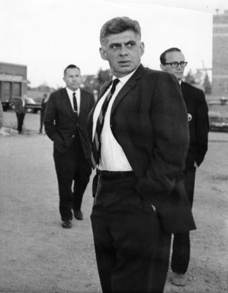 Jerry Wurf in foreground, hands in pockets, slightly turned and looking at someone out-of-frame; two figures stand at a distance behind him.