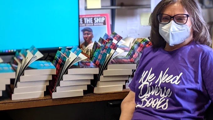 Karol Ruth Silverstein is sitting in a desk chair in front of a counter filled with stacks of the CURSED paperback, ready to be signed. 