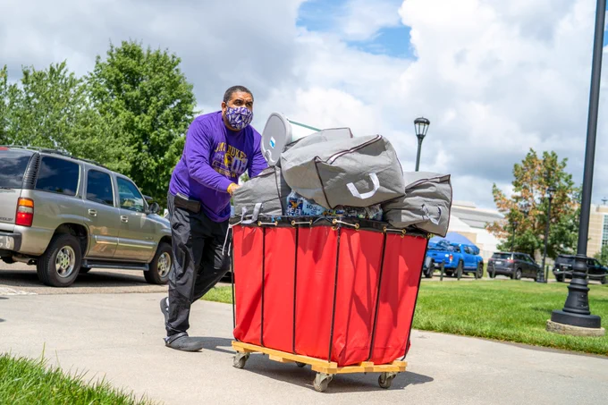 A volunteer wearing JMU shirt rolls full luggage dolly towards East Campus dorm.