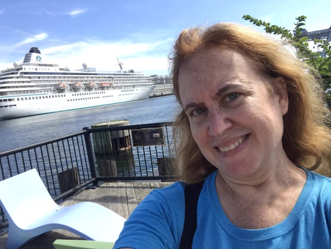 Lisa in the foreground smiles at the camera while in the background is a starboard side view of the cruise ship Crystal Symphony docked adjacent to Boston’s Design Center.