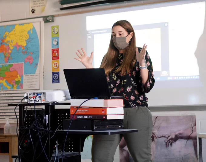 Teacher stands at front of classroom teaching with excited hands outreached.