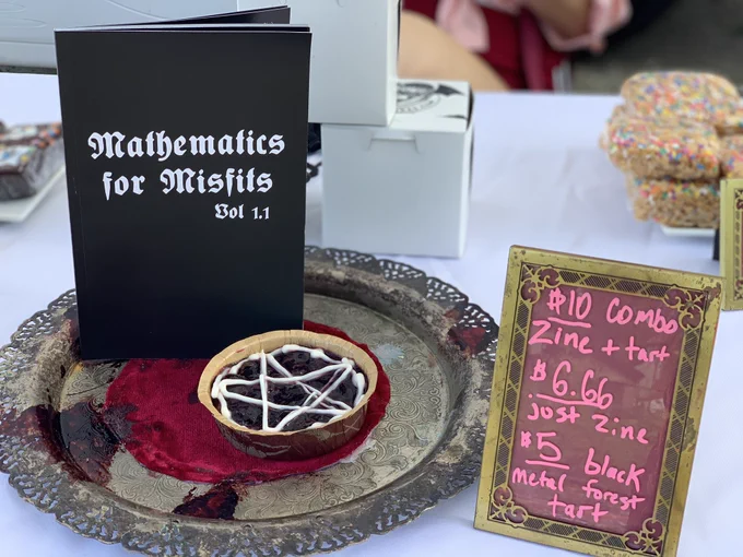 Mathematics for Misfits book standing behind a mini Black Forest tart with white chocolate ganache pentagram topping, both in an ornate golden dish. To the right is a small sign displaying the prices: $10 combo, $6.66 for the book alone, and $5 for the tart alone.