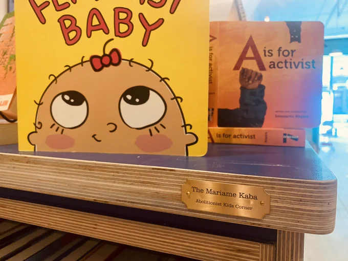 Shot from below, the plaque “The Mariame Kaba Abolitionist Kids Corner” under the board books Feminist Baby and A is for Activist