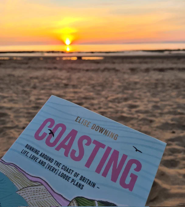 Photo shows a bright orange sunset over the sea. The top of the book is show in front of the sand and sea, with the title 'Coasting' visible
