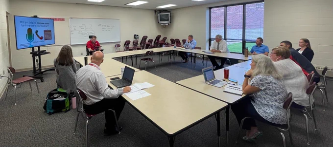 A classroom with a large square shaped table configuration.  Teachers/Administrators surrounding the tables.  The presenter, AccessJer20 is at the front of the room, presenting from an iPad with a Large TV.  