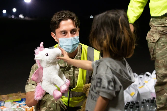 Male soldier in hi-vis vest and mask holds up a toy unicorn to a child.