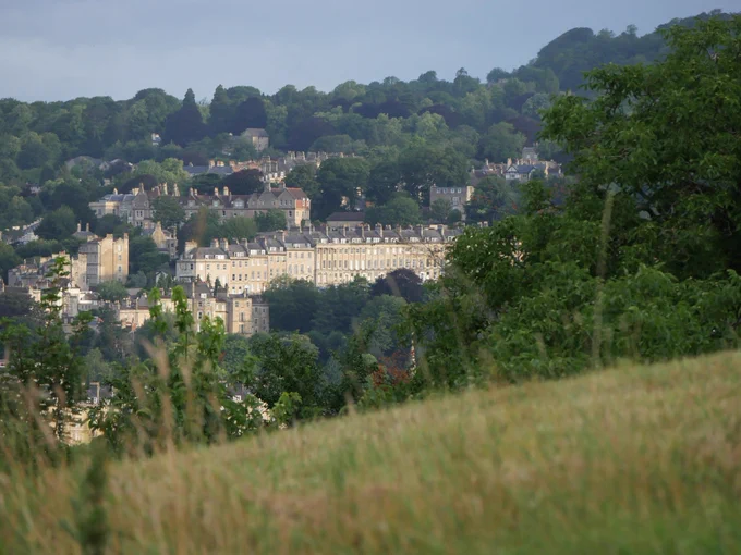 The lower half of a crescent of Georgian buildings with trees beyond and trees and a strip of meadow in the foreground.