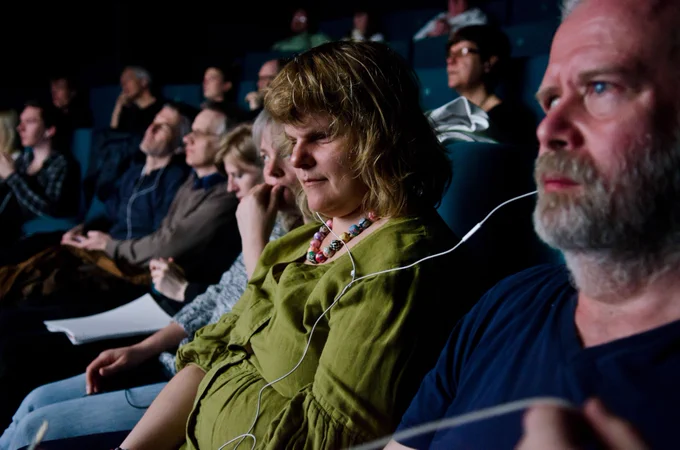 2 people who are blind sitting in a theater audience with listening devices providing audio description of the performance. 