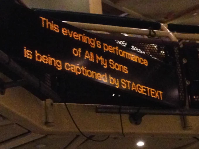 A screen in a theater that reads This evening's performance of All My Sons is being captioned by stagetext.