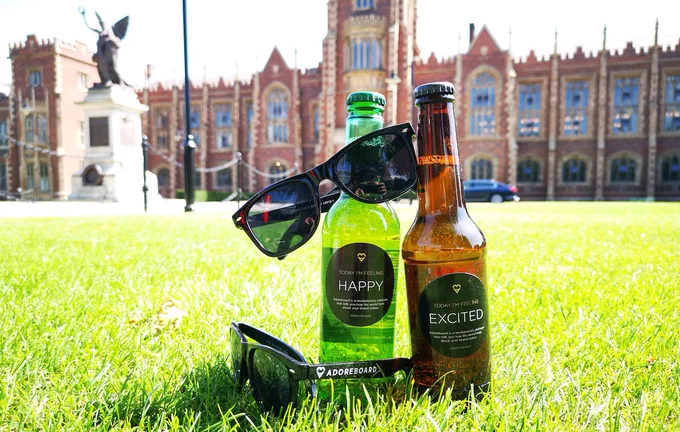 Image shows the Lanyon building with Adoreboard sunglasses and bottles on the grass. The labels on the bottles say Today I'm Feeling Excited and Happy.
