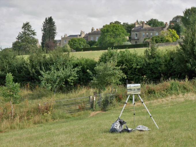 An easel set up in a field with a view of distant buildings, hedges and an orchard.