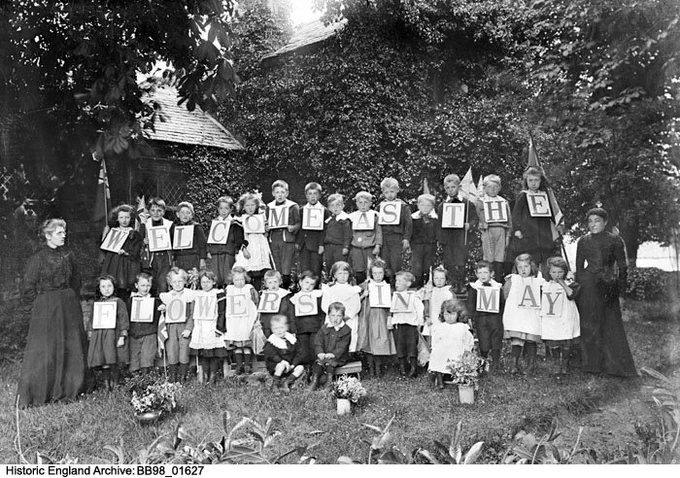 Black and white photo - Surrounded by trees and foliage, 2 rows of children in Victorian clothes with a lady in dark clothes stood at each end. The children have signs hung around their necks spelling out "WELCOME AS THE FLOWERS IN MAY".