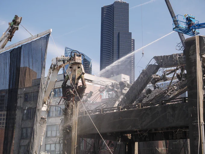 Machinery demolishing a concrete highway while crew members spray the work area with hoses to contain dust