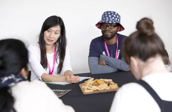 Sandy and Nolan are sat at a table, both listening in conversation to someone off-camera. Sandy is wearing a white top. Nolan is wearing a dark t-shirt with a grey shirt underneath, and a bucket hat.