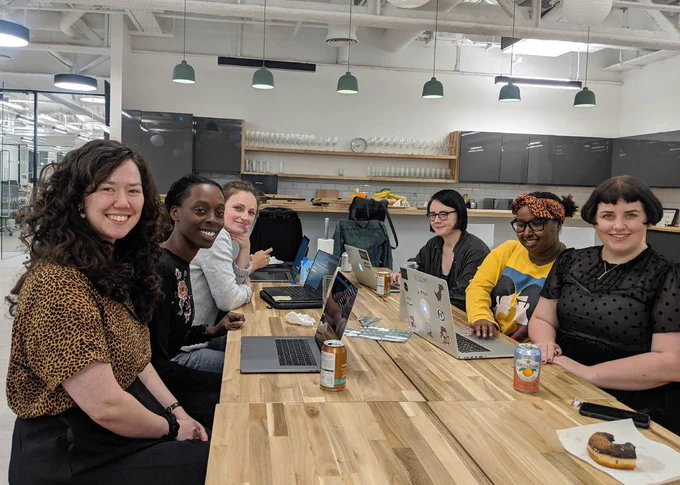 Six women at a picnic table with laptops, smiling at the camera