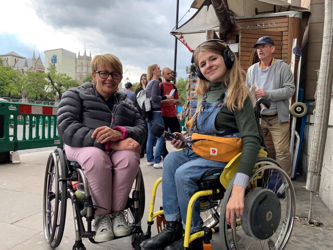 A photograph of Tanni and Katie smiling for the camera. Katie is wearing headphones and holding a microphone towards Tanni.