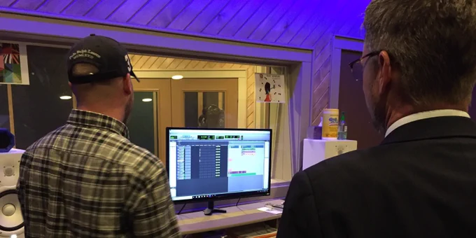Johnny Collett speaks to a Plaza Academy staff member in the Music Production Studio. There’s a studio room with glass in the back and computer with music editing software displayed on the screen