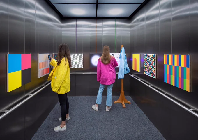 two visitors wearing brightly colored jackets and using ipods in an elevator with colorful panels hanging on the wall