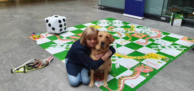 Ike, dark golden lab and dual purpose working dog looking straight into camera whilst being cuddled by his owner, Jo-Ann.  She has blond hair and wearing blue shirt with jeans.  They are both sitting on a garden sized board game with a blow up dice in the background.