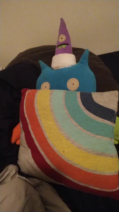 Blue felt bat stuffed animal hiding behind a rainbow pillow