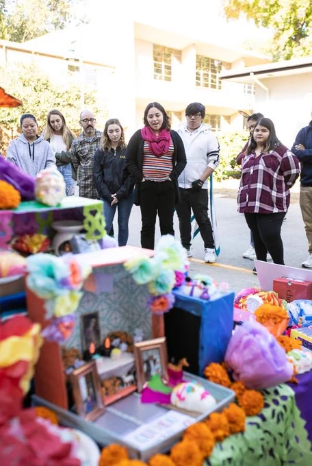 A group of students stands around a colorful alter with photos of deceased loved ones on it. One student is speaking as if to explain the alter to the others.