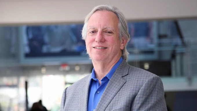 A man standing in an office wearing a grey suit and blue shirt smiles at the camera.