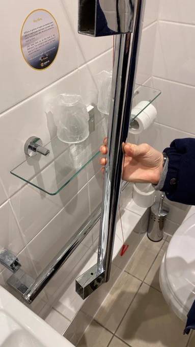 Jess Thom's PA holds up a posh, polished silver grab rail that is not attached to the wall. This is moments after it came away during a toilet to wheelchair transfer. A bathroom shelf and toilet roll are visible in the background.  