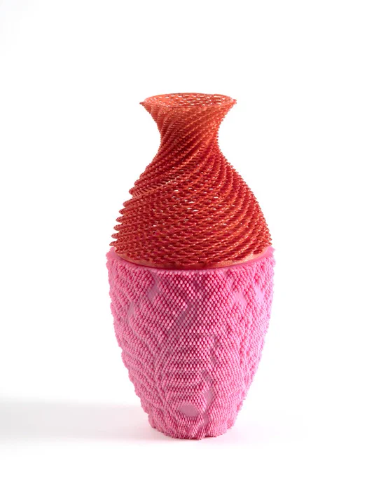 This is a tall narrow vase. The top of the vase is red and the bottom of the vase is bubble gum pink. The surface of the vessel has an intricate, tactile pattern. On the bottom of the vase, the pattern is in the form of large leaves or paisleys