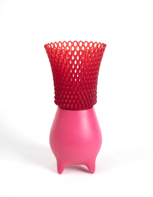 This is a suer modern vessel or vase. The top of the vase is an upside down cone form composed of red loops of plastic. The bottom of the vessel looks is smooth and bright pink. The bottom of the vessel has a cute blobby form with little legs.