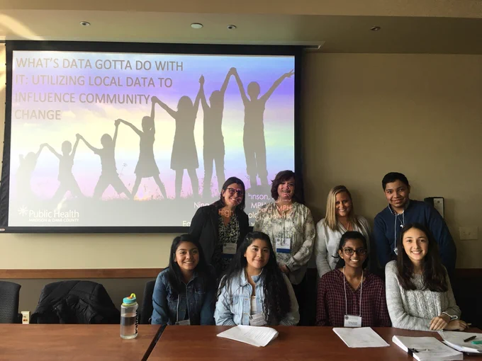 5 students and 3 Public Health professionals are lined up behind a desk. 4 young women are seated, and three women and one young man are standing behind them. In the background is a screen with the presentation title in black letters over an image of silhouettes over a sunset.