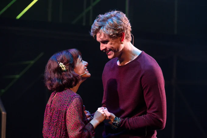  Photo credit: Leslie Schachter Hades and Persephone look lovingly at each other and hold hands. Hades wears a burgundy sweater and has blonde curly hair. Persephone wears a patterned red and black dress and looks up at Hades. She has shoulder length brown hair with a flower pin.