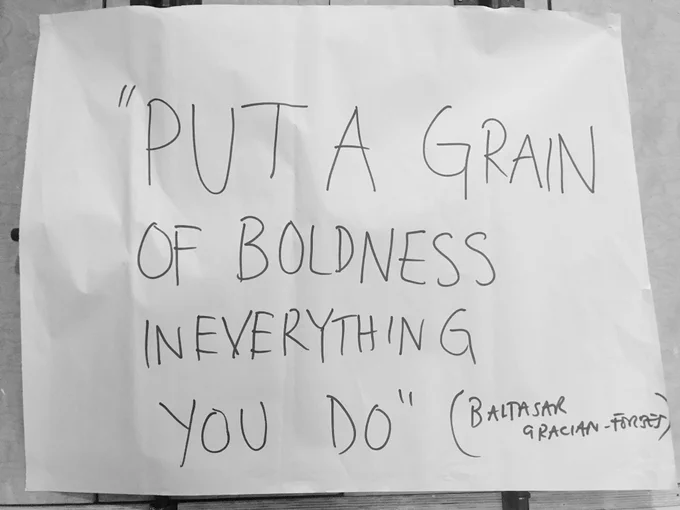Black & white photo: White paper with scribbled quote in black ink saying “Put a grain of boldness in everything you do” (Baltasar Gracian-Forest)
