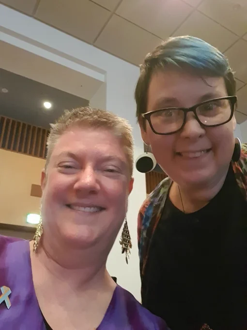 2 people smiling, 1 has blue hair, the other is wearing purple, both have impressive earrings