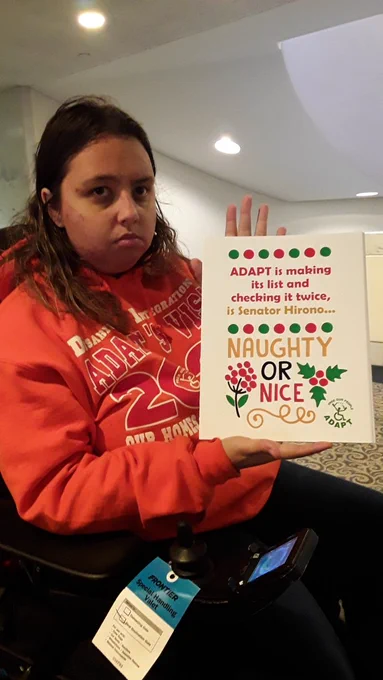 An ADAPT organizer sits in her wheelchair wearing a bright orange sweatshirt holding a holiday card that reads "ADAPT is making its list and Checking it twice is Senator Hirono Naughty or Nice?" With an ADAPT Free our people logo in the bottom right corner.
