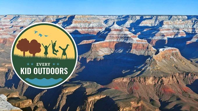 Image description: a posterized logo showing 3 silhouetted children playing in a grassy meadow with 2 trees. In the foreground a pool of water. Text reads: "Every Kid Outdoors." Logo is superimposed above a background of snow covering peaks and cliffs within a canyon landscape.