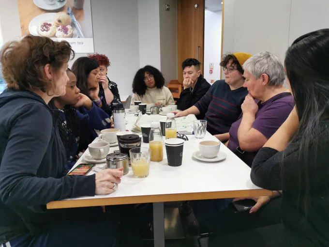 9 very thoughtful looking people sat around a table covered in cups of coffee and glasses of orange juice