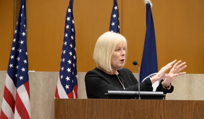 Director General Carol Perez indoors at podium speaking with American flags in the background