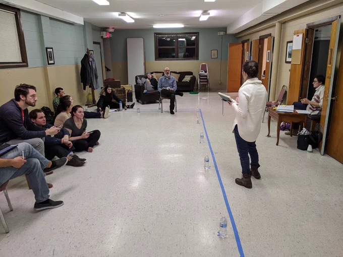 Woman lecturing in a community center with 11 people sitting in various ways (floor, chair, etc.) listening intently.