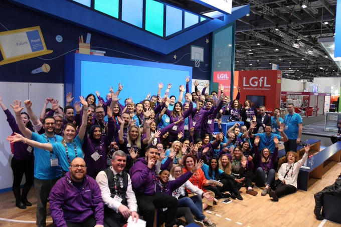 A group photograph of the Microsoft Education team at the Bett Education show at London ExCeL. There are many people of all different nationalities all wearing Microsoft education clothing. They are cheering and smiling towards the camera.