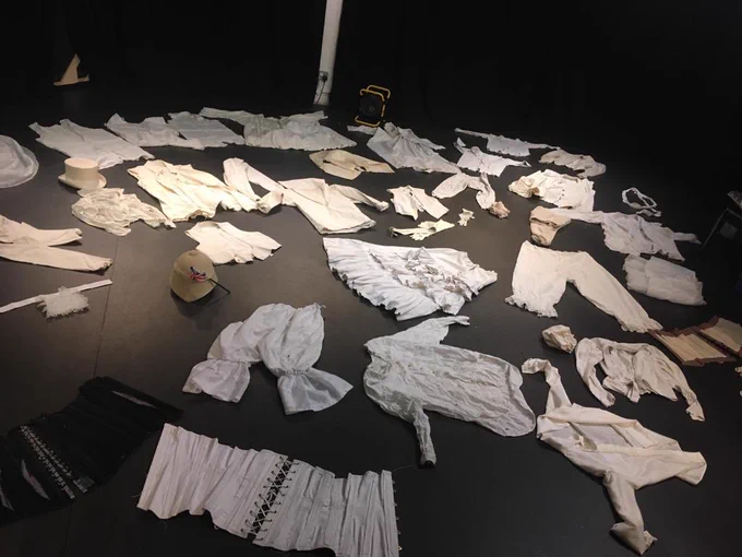 A variety of Victorian bits of clothing - corsets, shirts, long johns, waistcoats, hats, ties and gloves - spread out across a black floor.