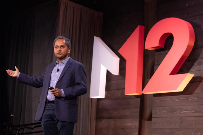 A man waering a suit stands on stage in front of a red and white M12 logo