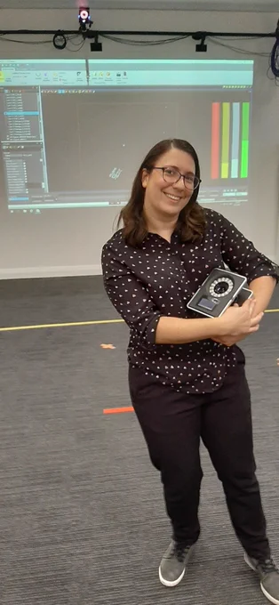 Photo of Jess smiling while cradling a large square camera in her arms. In the background, a specialised studio setup with cameras and projection on the wall.