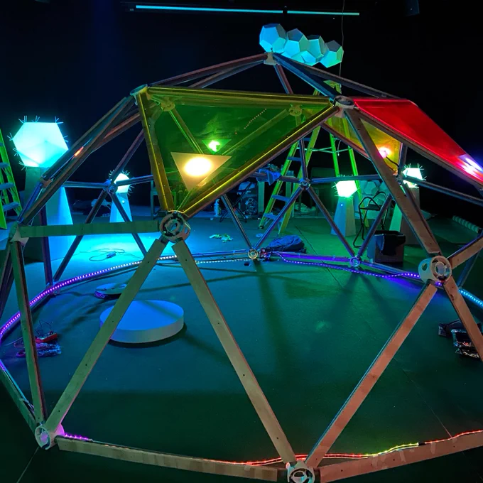 Photo of a geodesic dome made of plywood, with transparent coloured acrylic triangles, in a dark room lit by multicoloured lights both on the dome and from objects around the room glowing.
