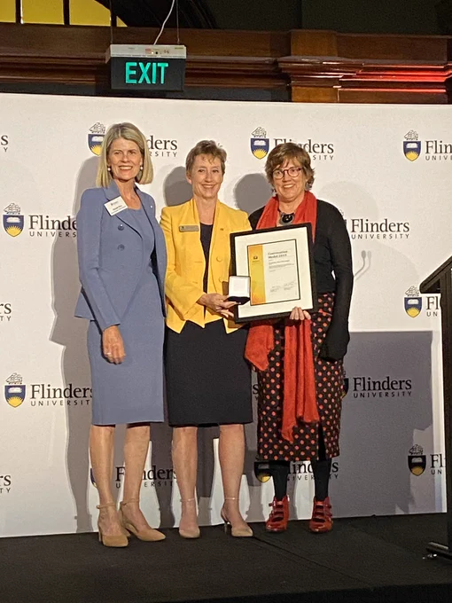 Three women standing on a stage. One on right holding framed award and medal.