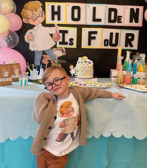 Holden wearing a cardigan, a T-shirt with a cartoon scientist and glasses smiling and posing in front of a lab equipment and a sign that reads “Holden is Four”