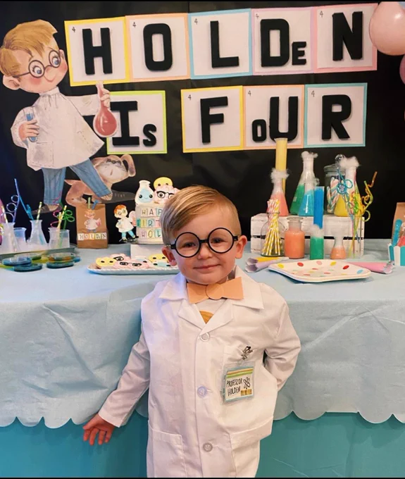 Holden wearing a lab coat, bow tie, and glasses standing in front of a lab equipment and a sign that reads “Holden is Four”